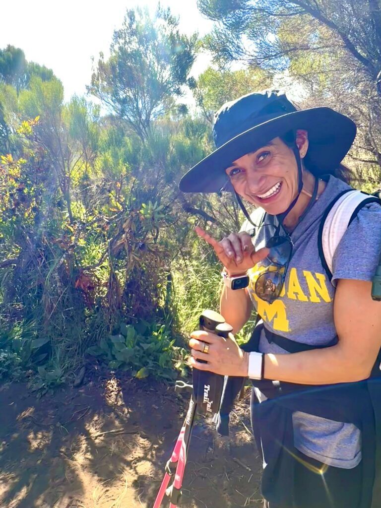 A woman wearing a hat and a t-shirt with the word can on it is hiking on a trail.