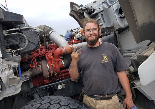 Man in a US Army shirt gives a rock-on hand gesture in front of a truck engine with open hood.
