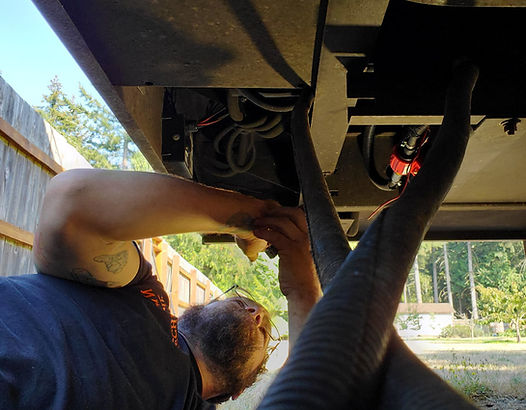 A person under an RV, working on hoses. Outdoors with a fence and a house in the background.