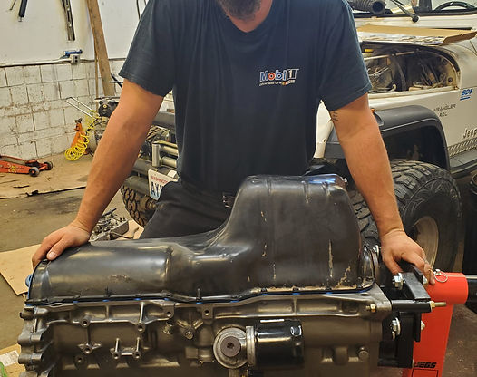 Man with hands on an engine, in a workshop setting. Dark shirt, vehicle visible in the background.