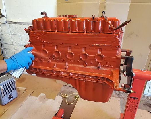 A person sprays orange paint on a car engine mounted on a red engine stand indoors.