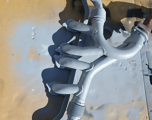 Gray-painted exhaust manifold on a cardboard surface, casting a long shadow in sunlight.