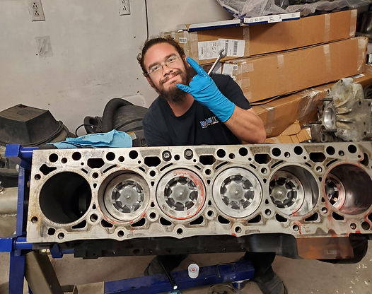 Man with blue gloves gives a rock-on hand gesture, stands behind an engine block with six visible cylinders.