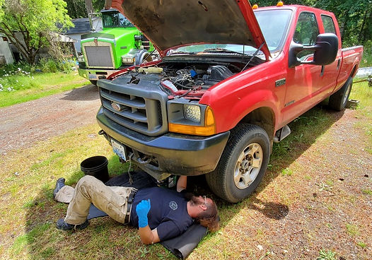 Man working on red truck, lying on mat. Hood open. Green truck in background.