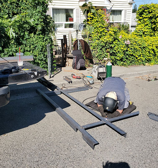 Person welding metal frame outdoors on pavement with a house in the background.