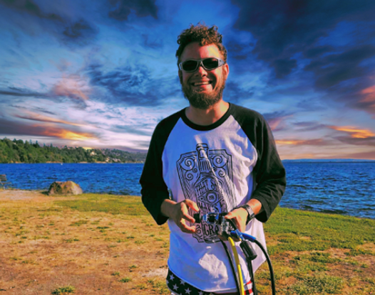 Man in sunglasses smiles at the camera on a beach; holding equipment, blue water, sunset sky.