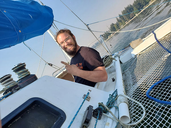 Man on sailboat, smiling, pointing, sunny day. Boat is white with blue sails.