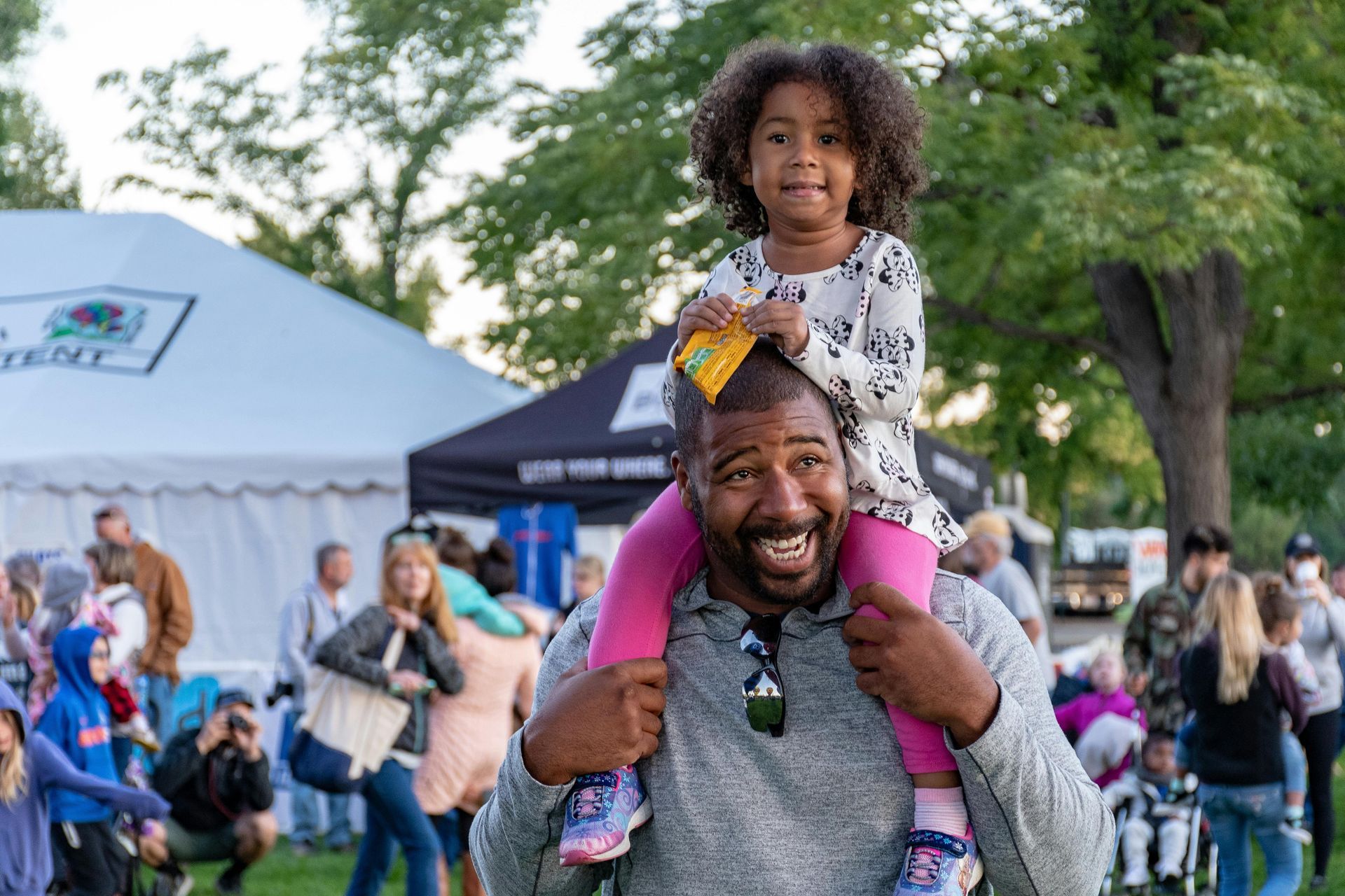 A man is carrying a little girl on his shoulders at a festival.