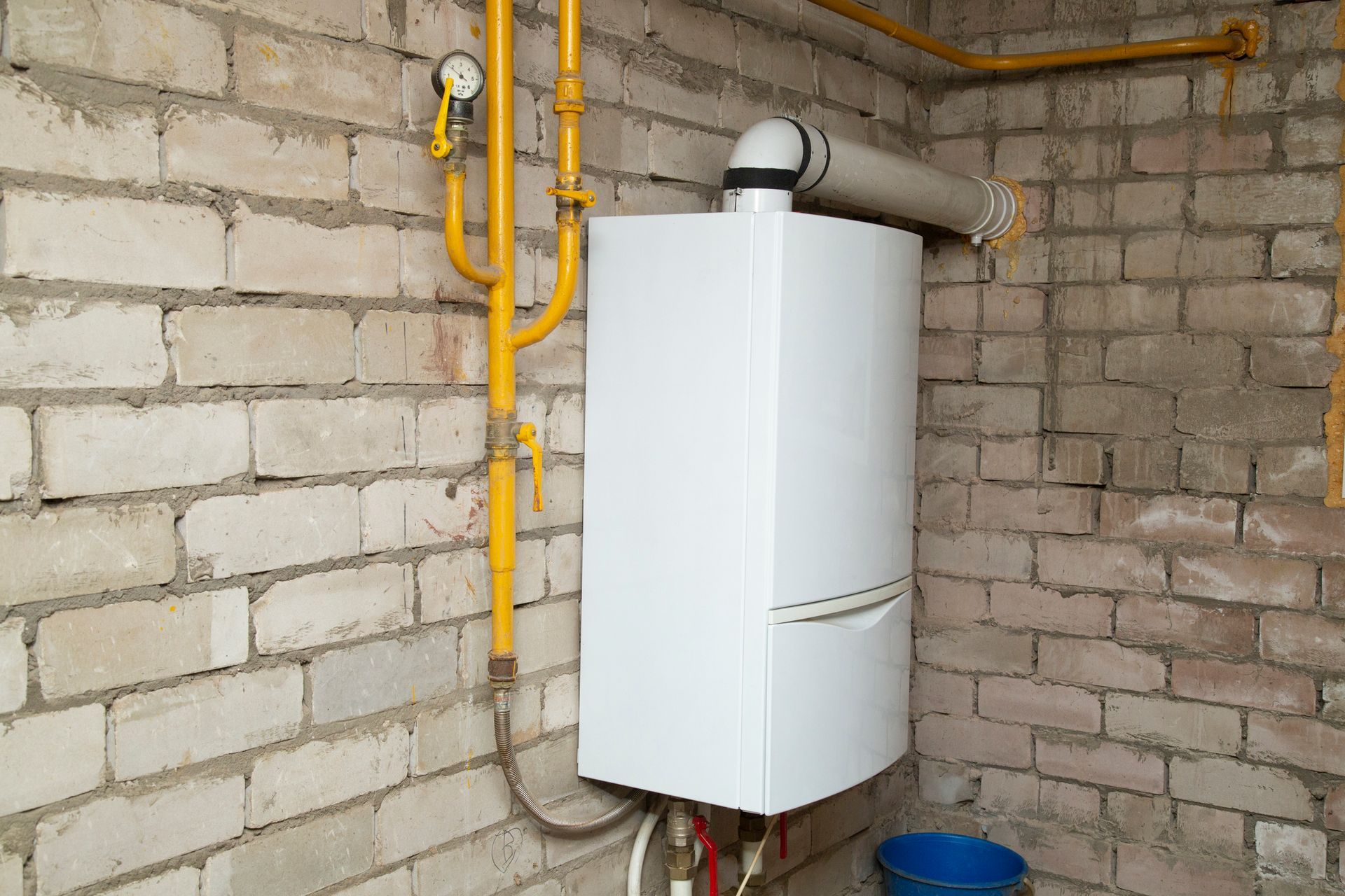 White gas furnace attached to a yellow gas pipe, against a brick wall.