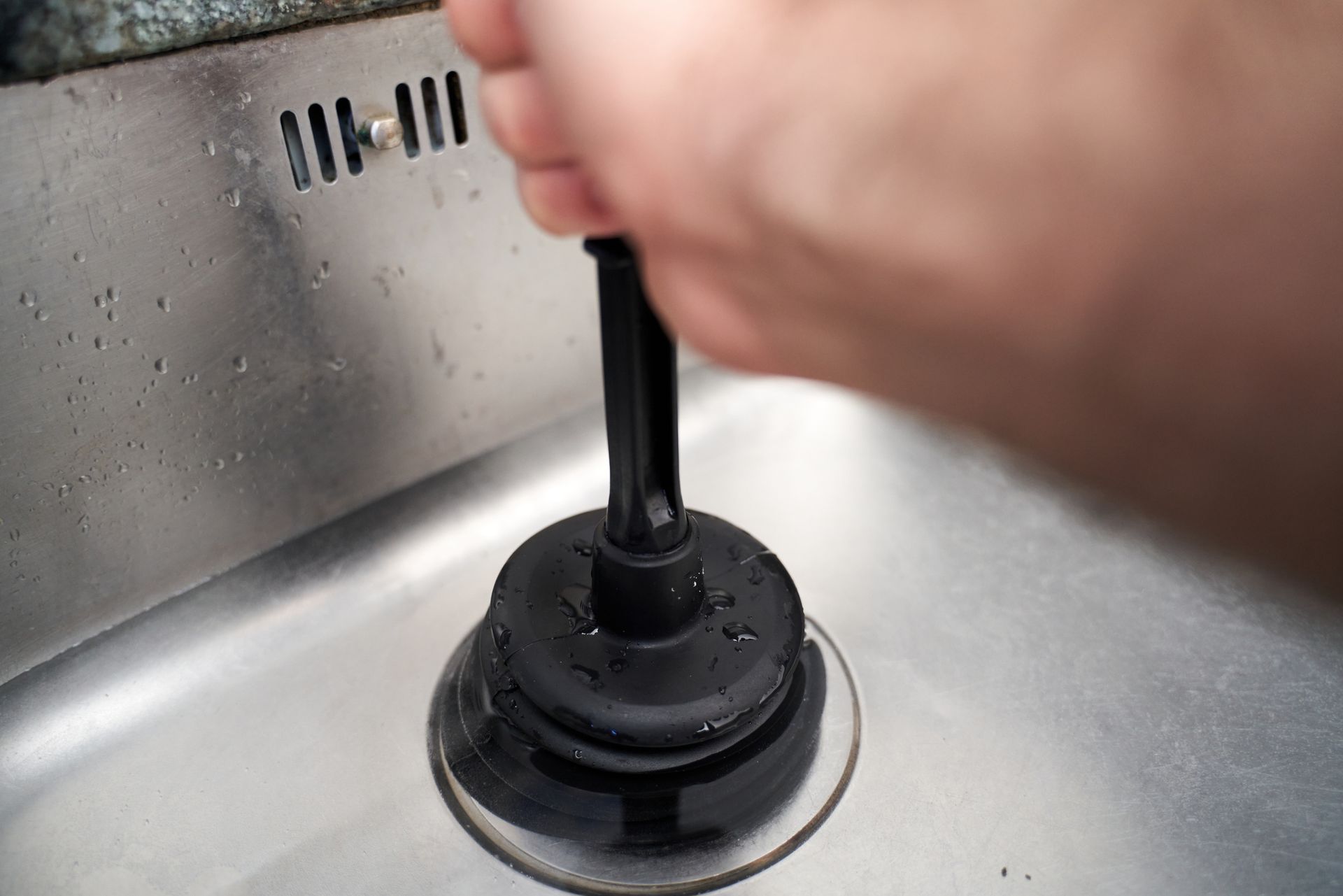 Hand using a black plunger to unclog a stainless steel sink drain.