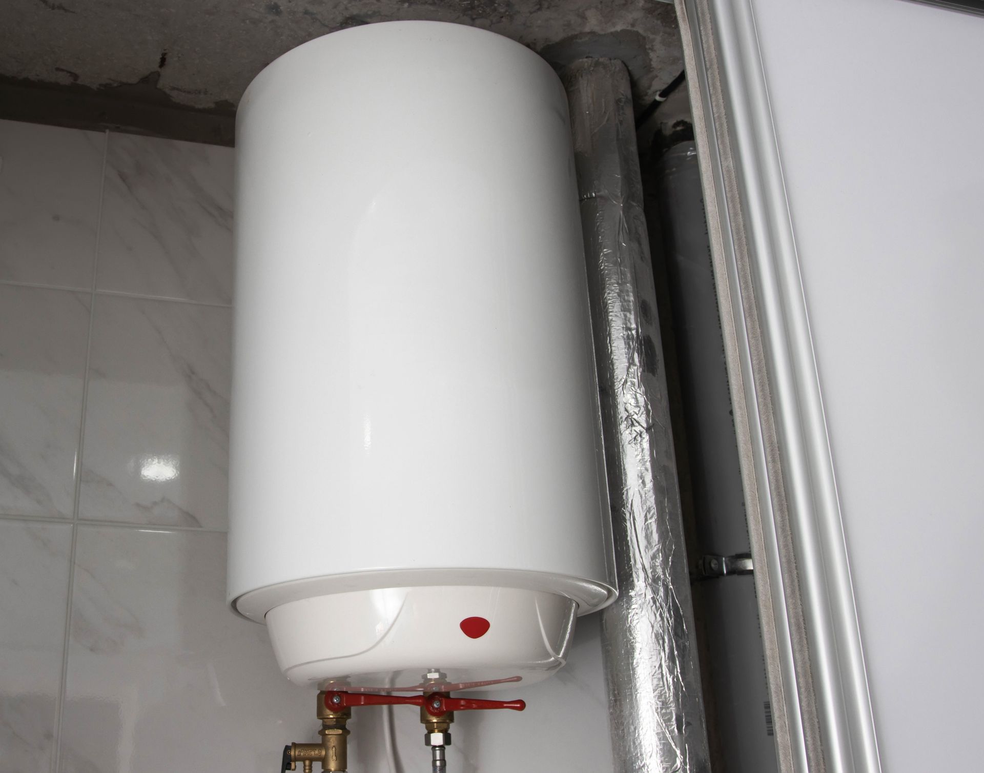 White water heater mounted on a tiled wall, with plumbing visible below.