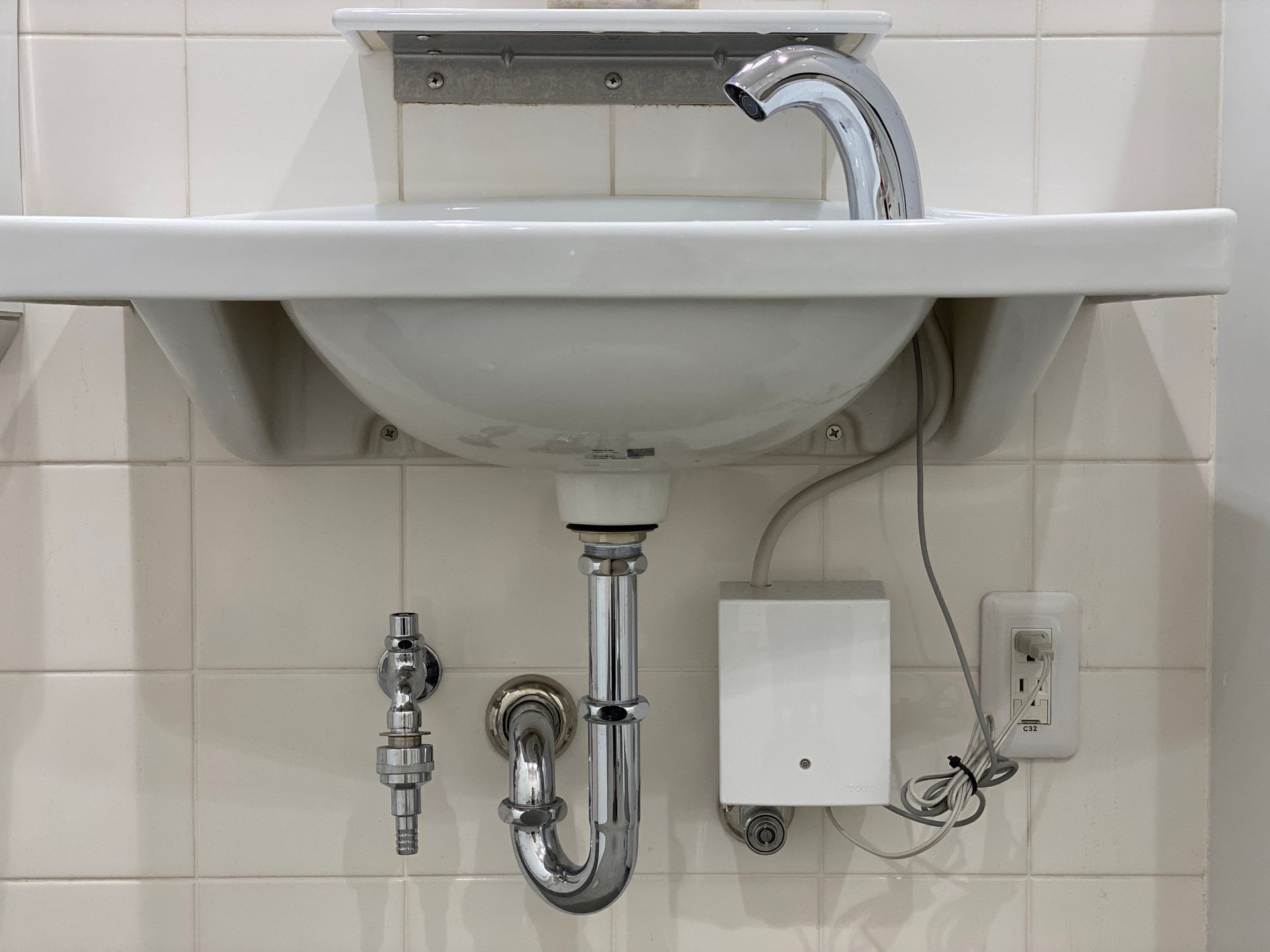 White sink with chrome faucet and plumbing against white-tiled wall, with a white electrical box and outlet.