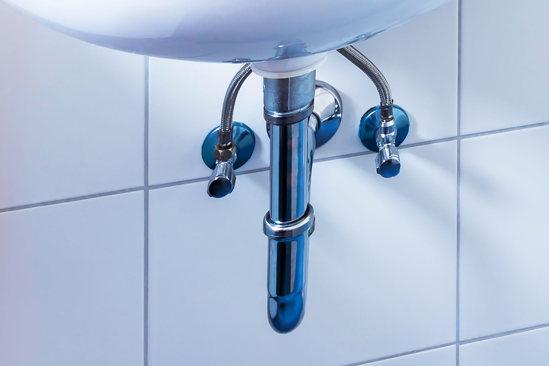 Underside of a white sink with chrome plumbing against white tiled wall.