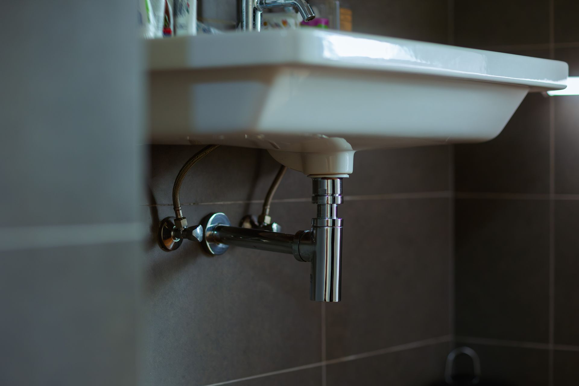 White bathroom sink with visible plumbing against a brown tiled wall.