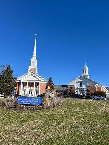 Two churches with spires, one brick and one white, under a clear blue sky. Rainbow flag displayed.