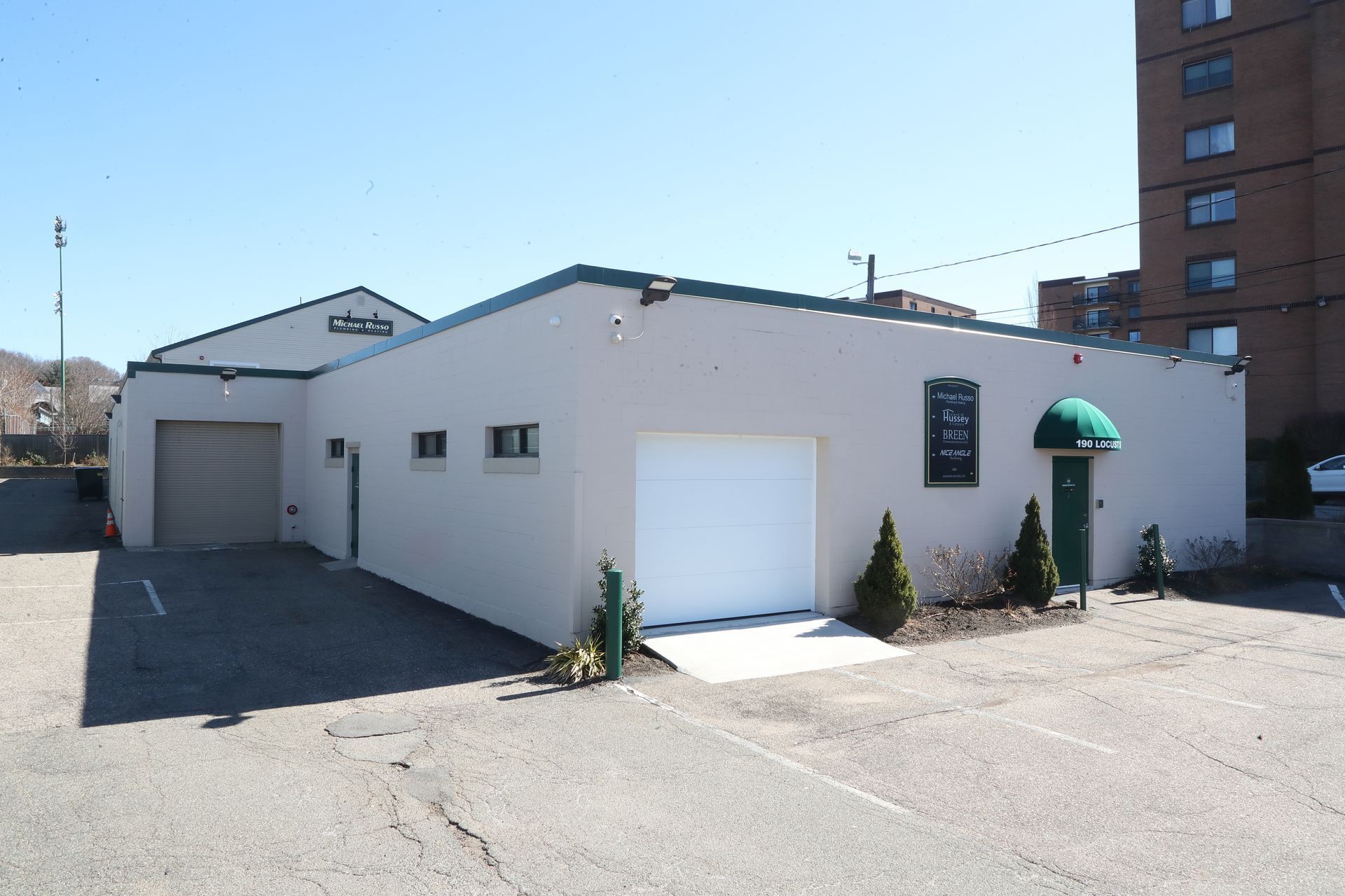 Tan building with garage doors, a green awning, and parking area on a sunny day.