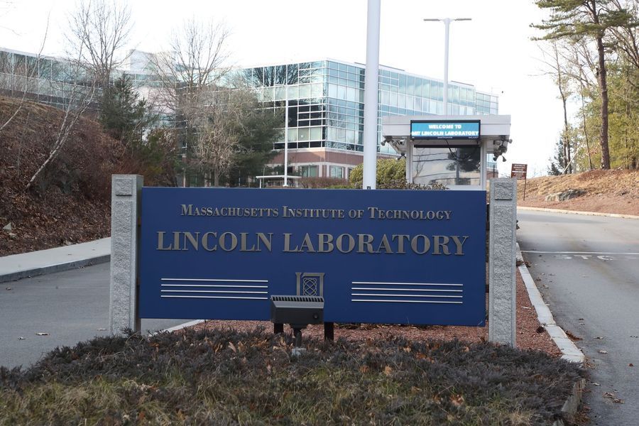 Sign for MIT Lincoln Laboratory, blue with gold lettering, building in background.