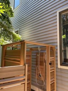 Wooden outdoor shower against a gray-sided house. Shower head and fixtures visible.