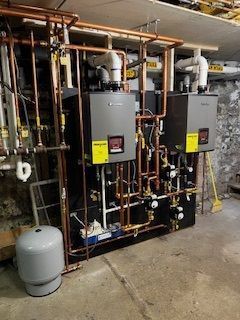 Two gray water heaters with copper pipes in a basement.