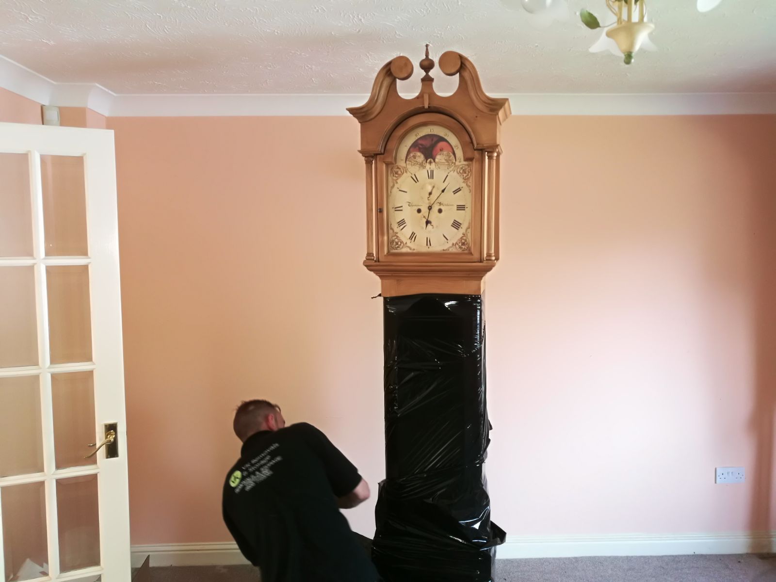 Man wrapping tall, antique clock in black plastic, in a room with pink walls and a white door.