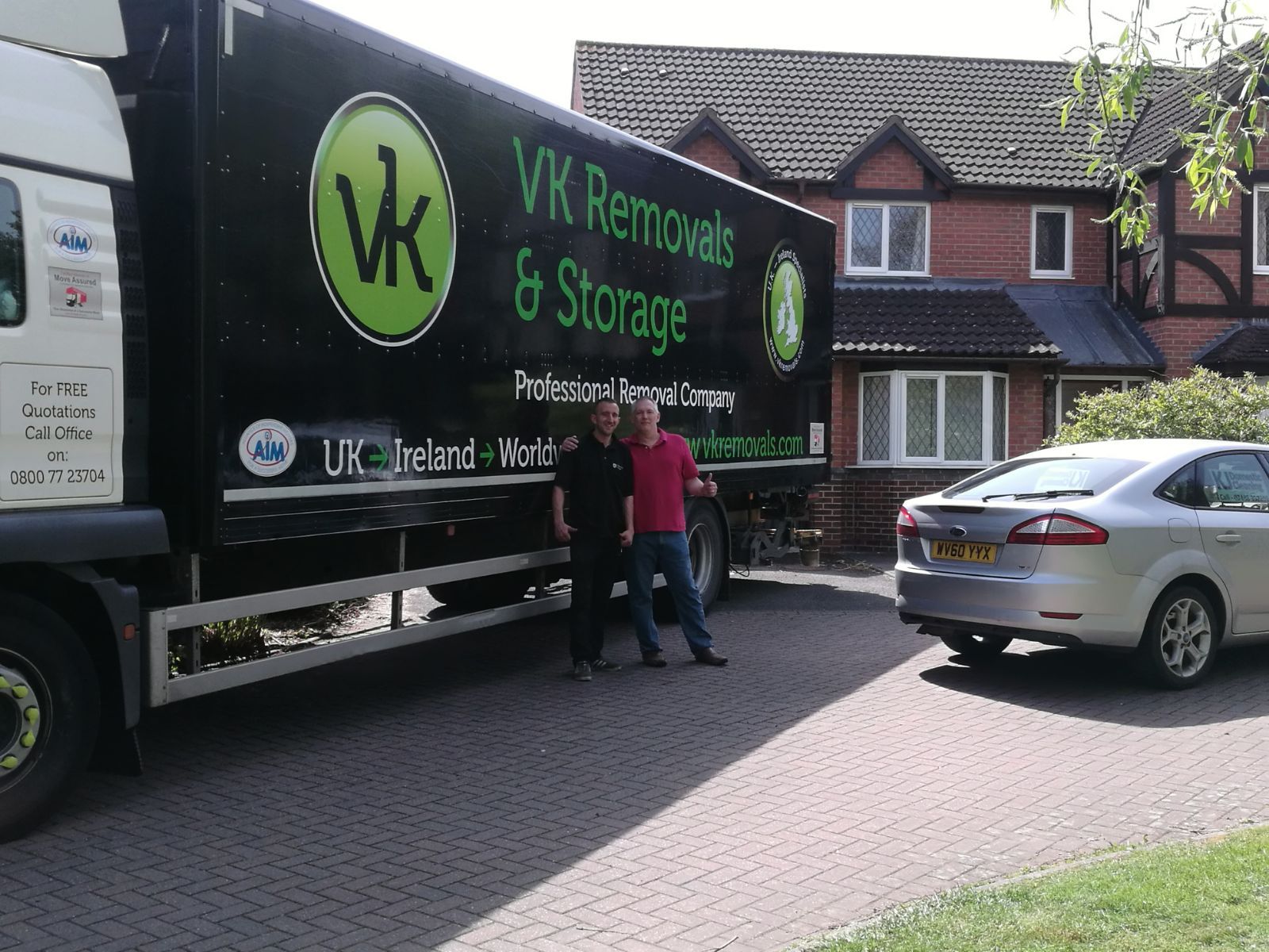 Two men by a VK Removals truck in front of a house with a gray car parked nearby.