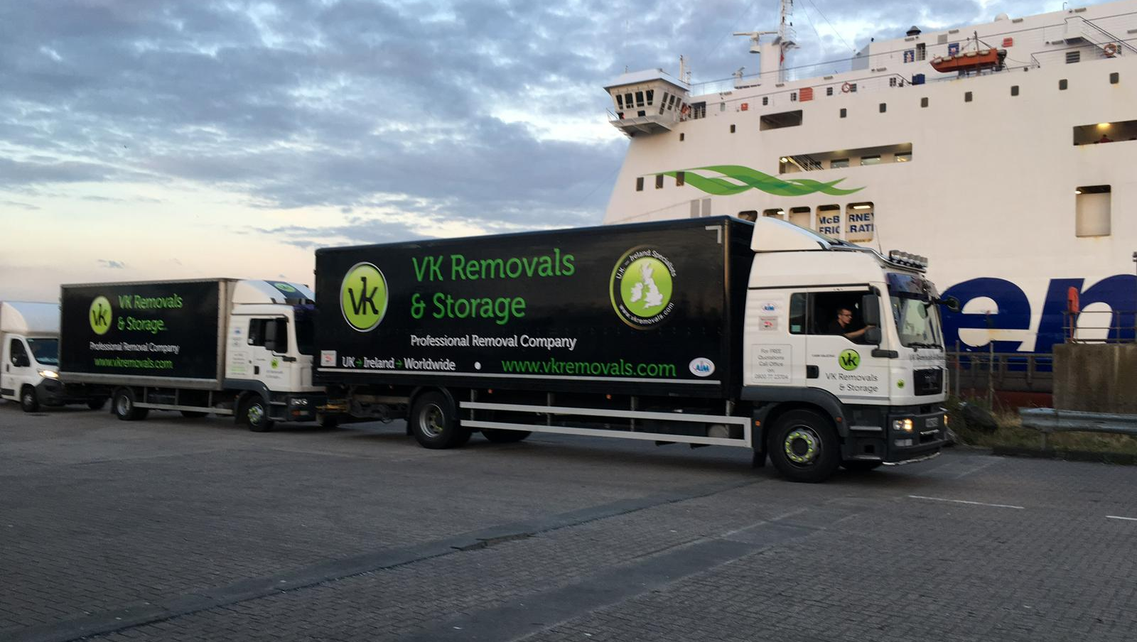 Two VK Removals trucks with logos parked near a ferry; sea in the background.