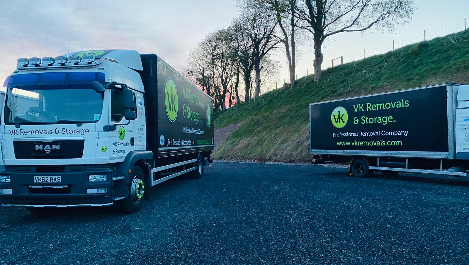 Two white and black removal trucks parked, with a green hillside in the background.