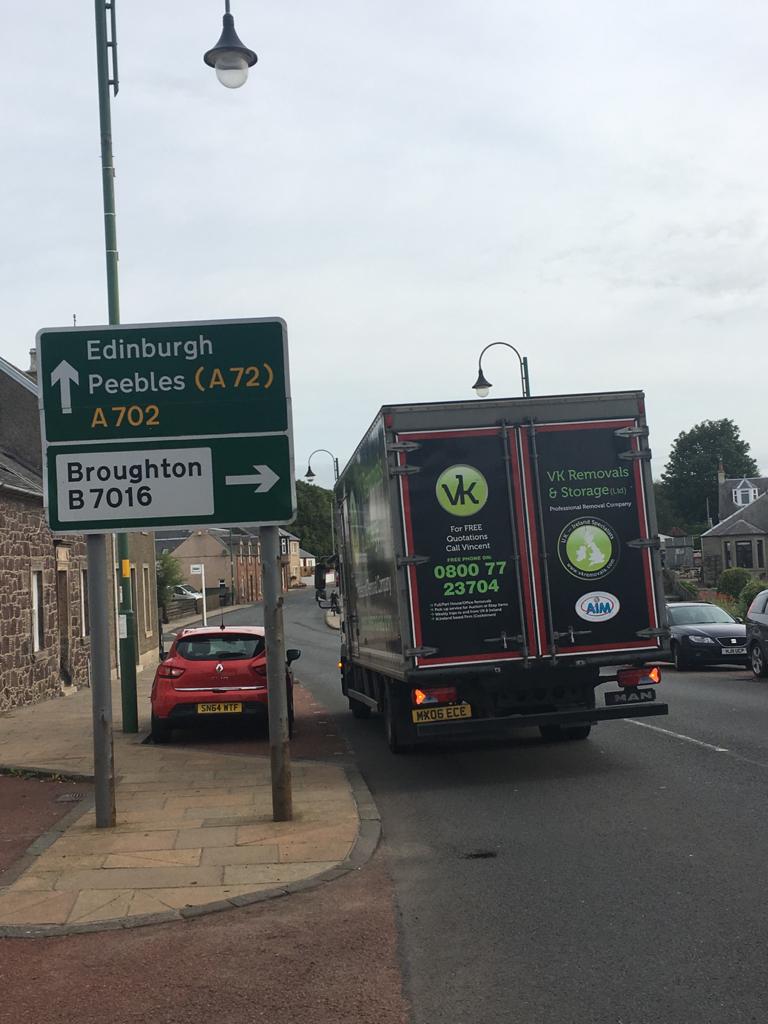 Signpost for Edinburgh and Peebles. A truck is parked on the street next to a parked red car.