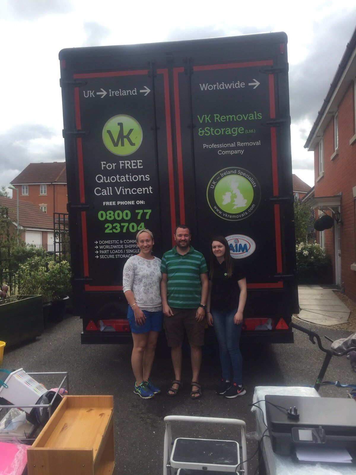 Three people pose in front of a black moving truck. The truck has company logos and contact info.