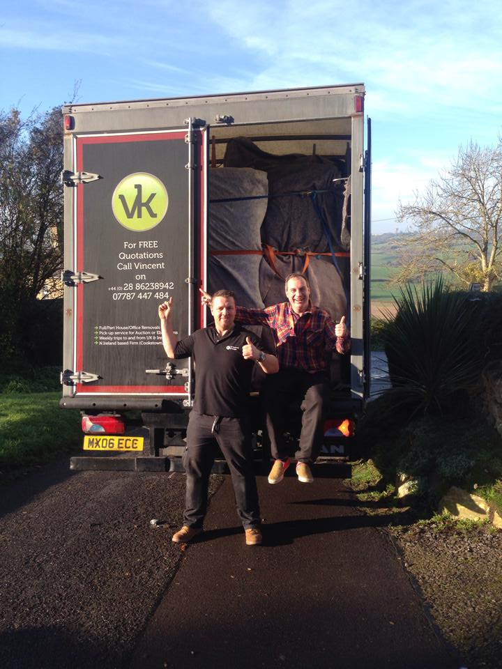 Two men by a moving truck, open doors revealing packed contents. One man raises his arms. Sunny outdoor setting.
