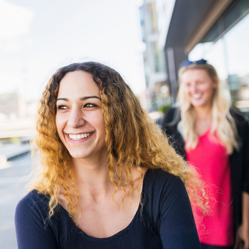 Smiling woman with curly hair outdoors; another woman smiles in background.