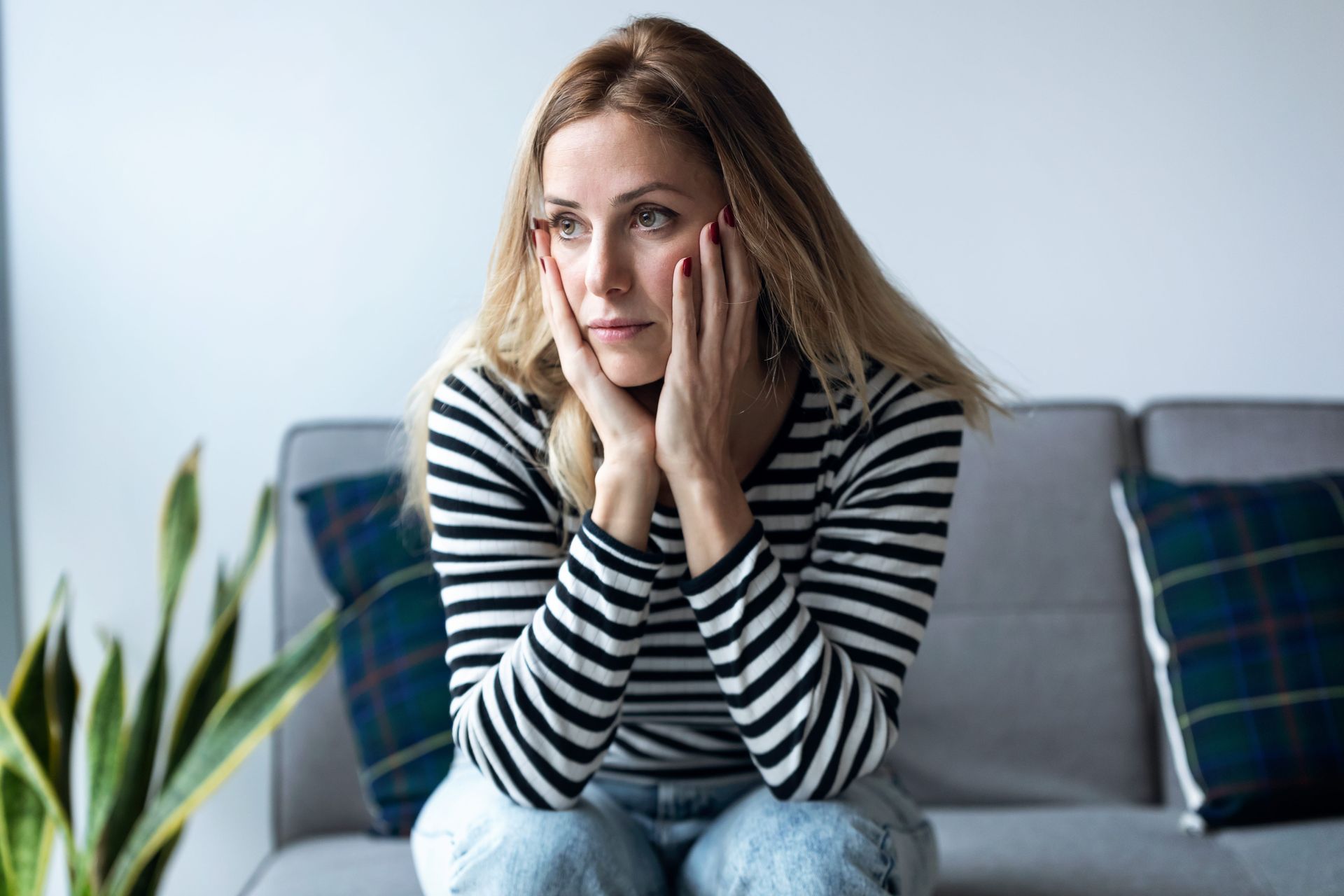 Woman with blonde hair, hands on cheeks, sitting on a sofa looking distressed.