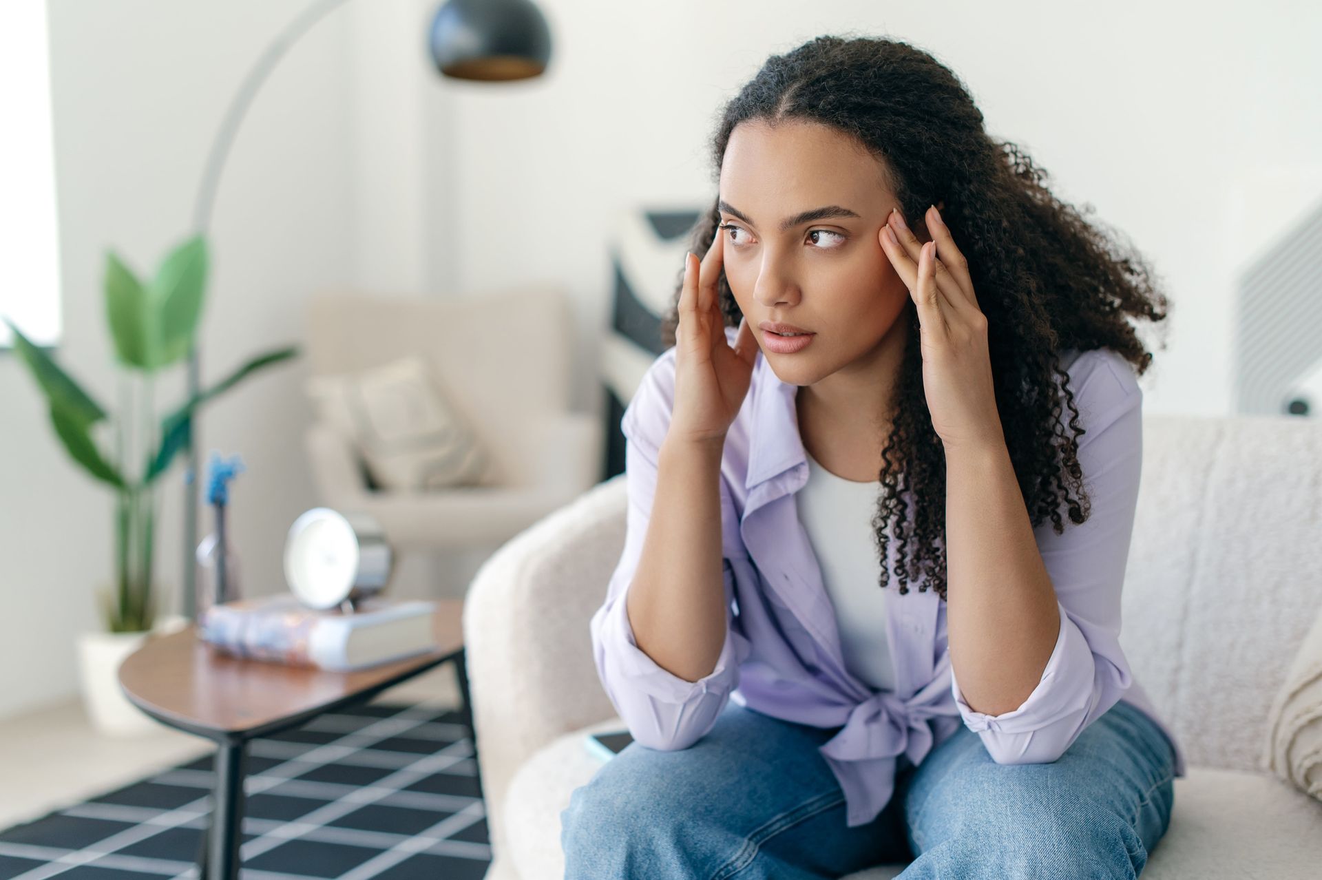 Woman with hands on temples, appearing stressed, seated on a couch in a living room.