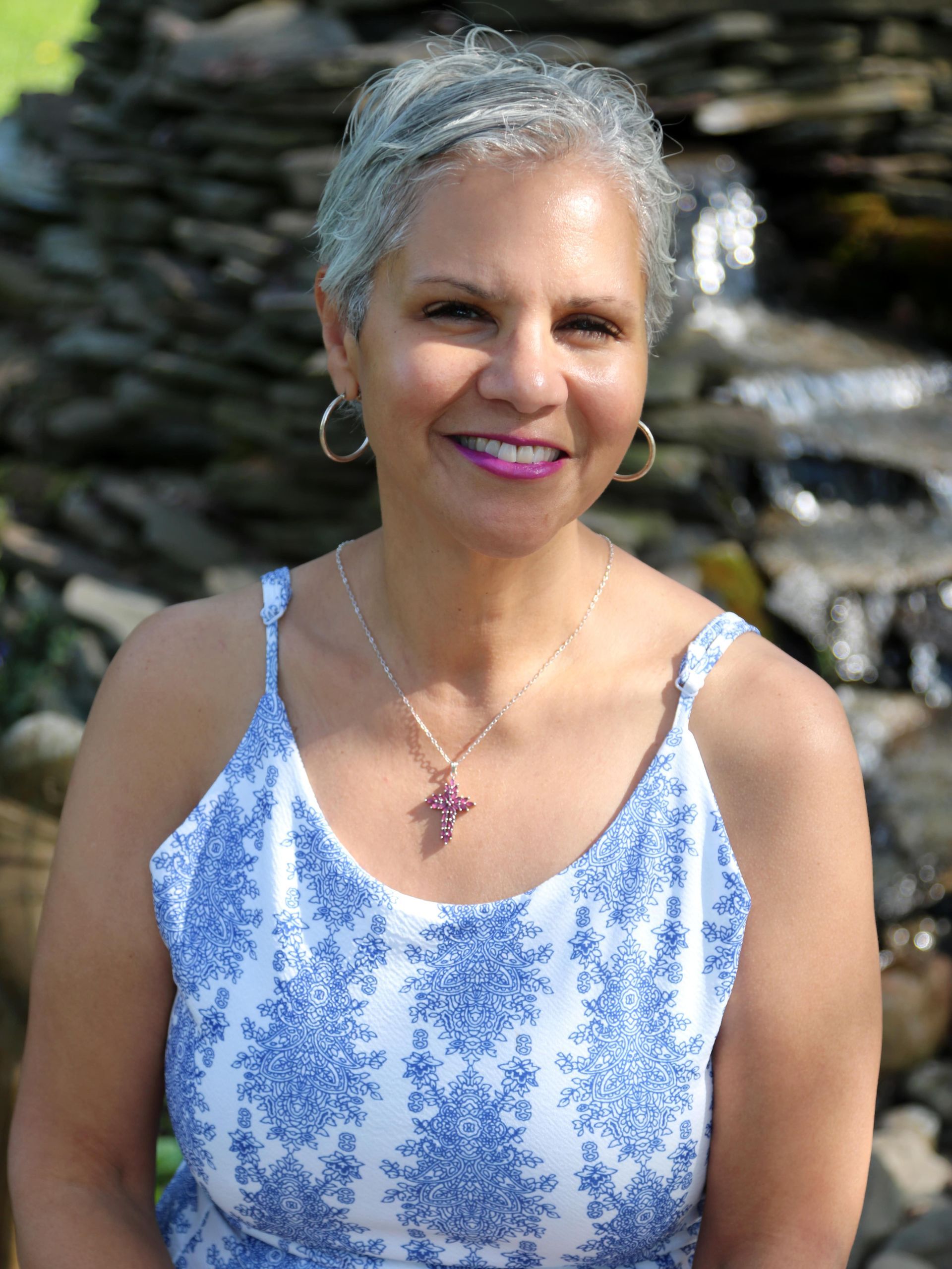 Woman with silver hair smiles, wearing a blue and white dress, and a cross necklace.