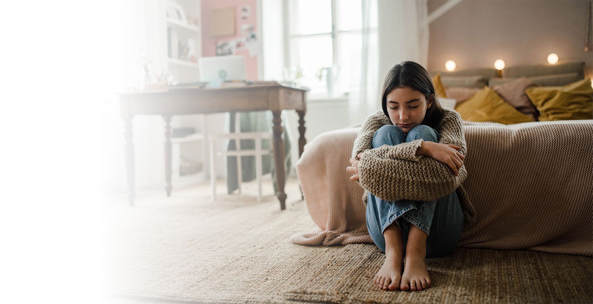 A young woman sits on a rug, hugging her knees, in a bedroom.