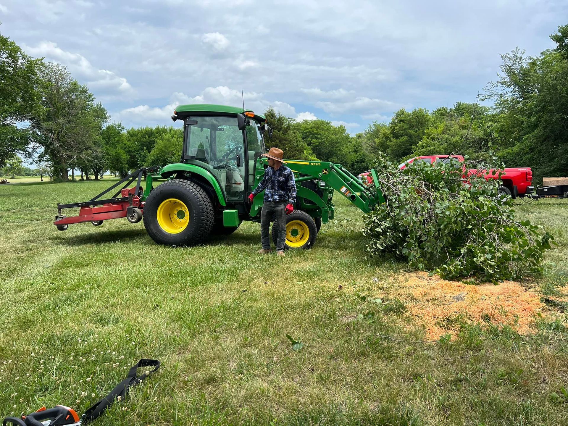 Man standing near a green tractor, loading branches in a field. Bright, sunny day.