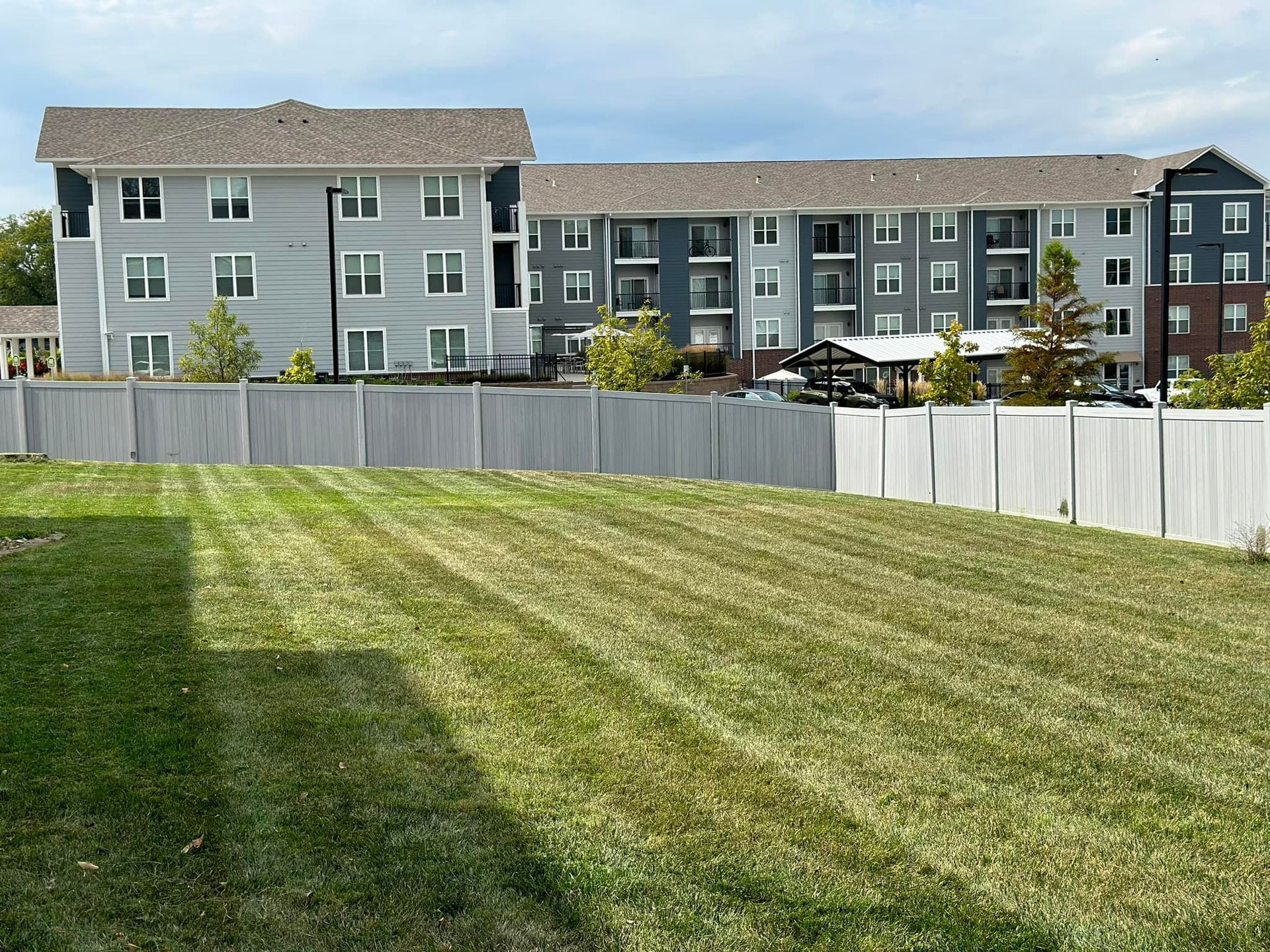 Well-kept lawn with a tall, white fence in front of a multi-story apartment building with gray and blue siding.