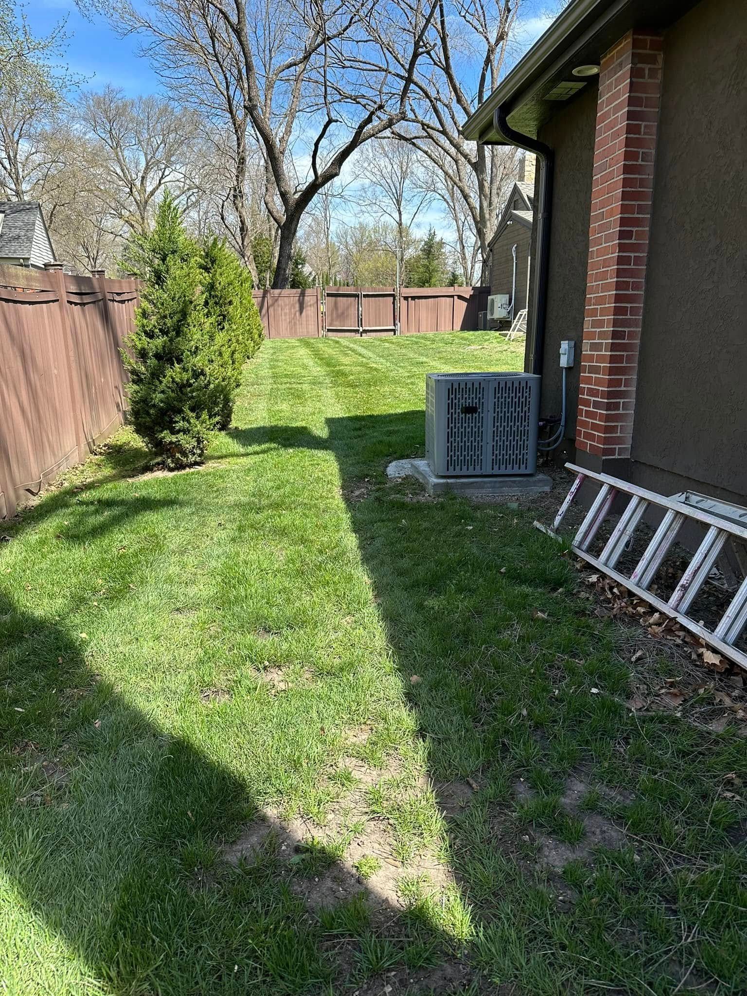 Lawn with a brown fence, green grass, a small tree, and a building with a brick pillar.