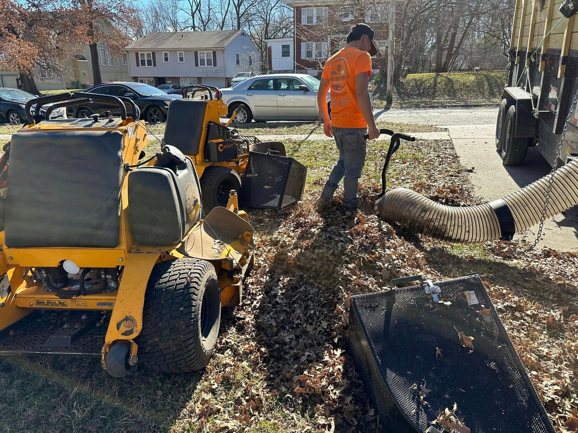 A worker uses a leaf vacuum to collect leaves next to a yellow lawnmower on a residential street.