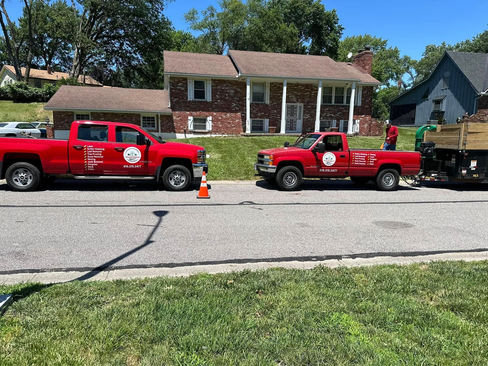 Two red pickup trucks parked on a street in front of a brick house with lawn.