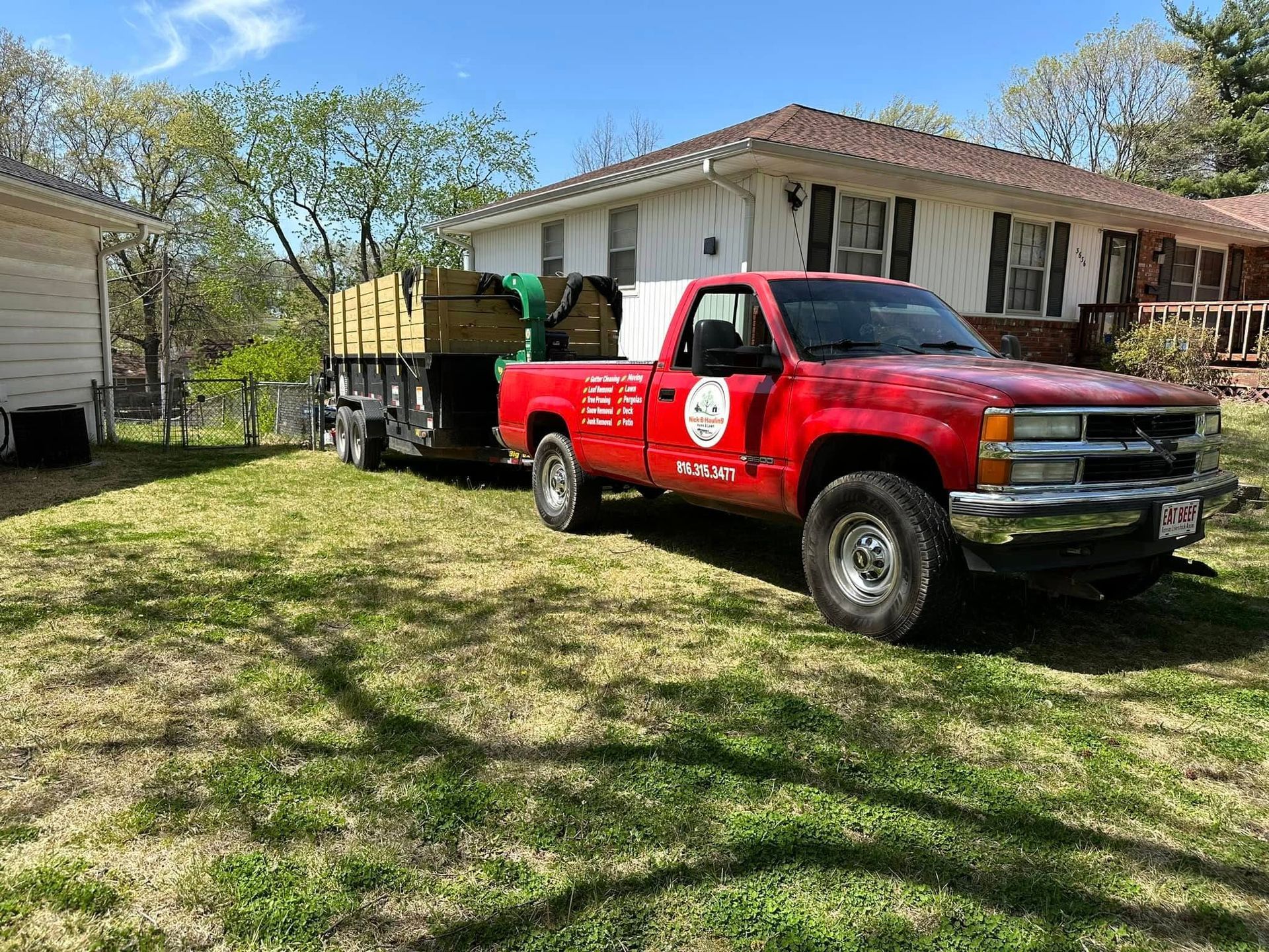 Red truck with trailer parked on grass, beside a white house; trailer holding lumber.