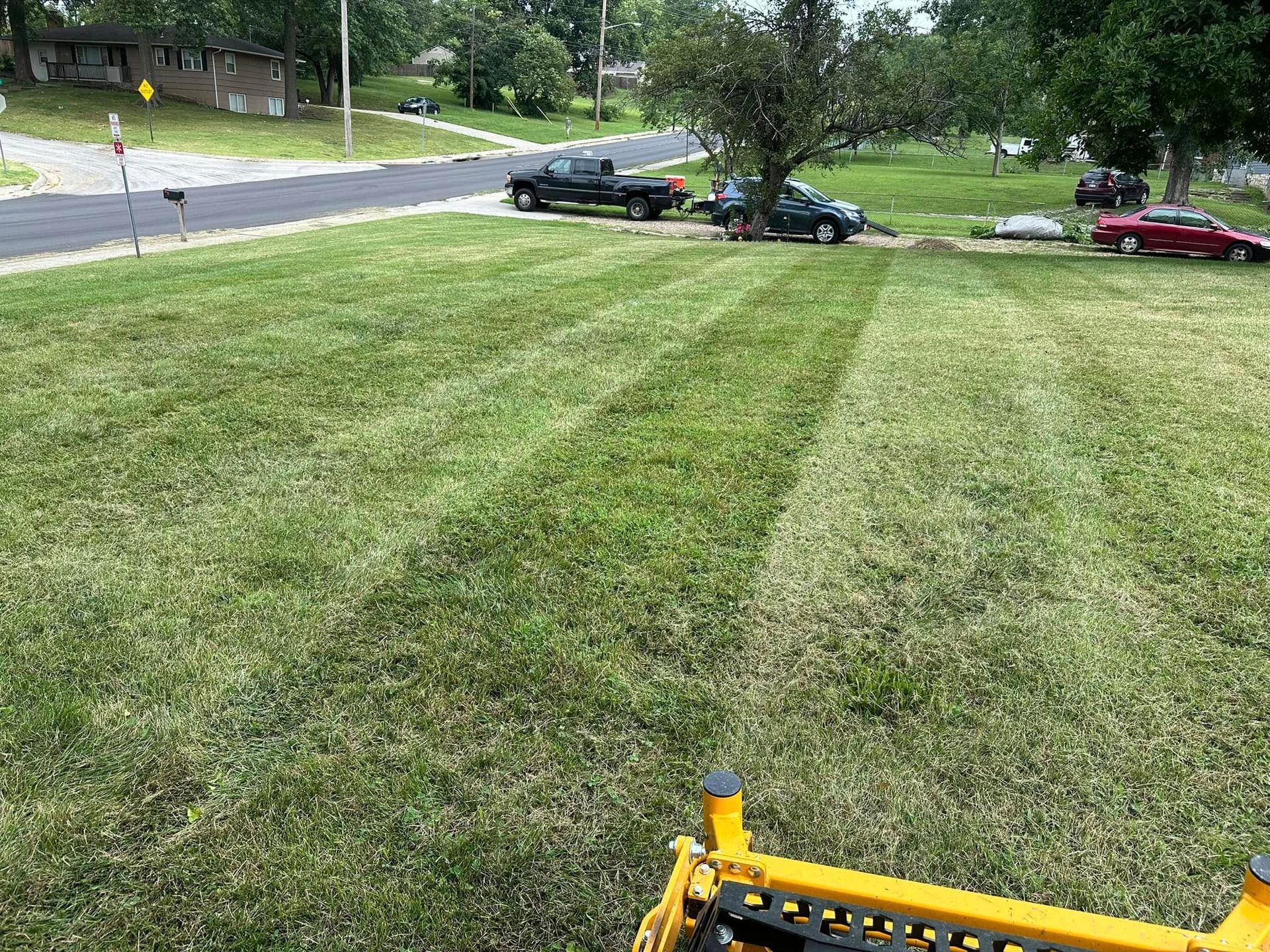 A lawn being mowed, showing freshly cut stripes in the green grass, with parked cars and a road in the background.