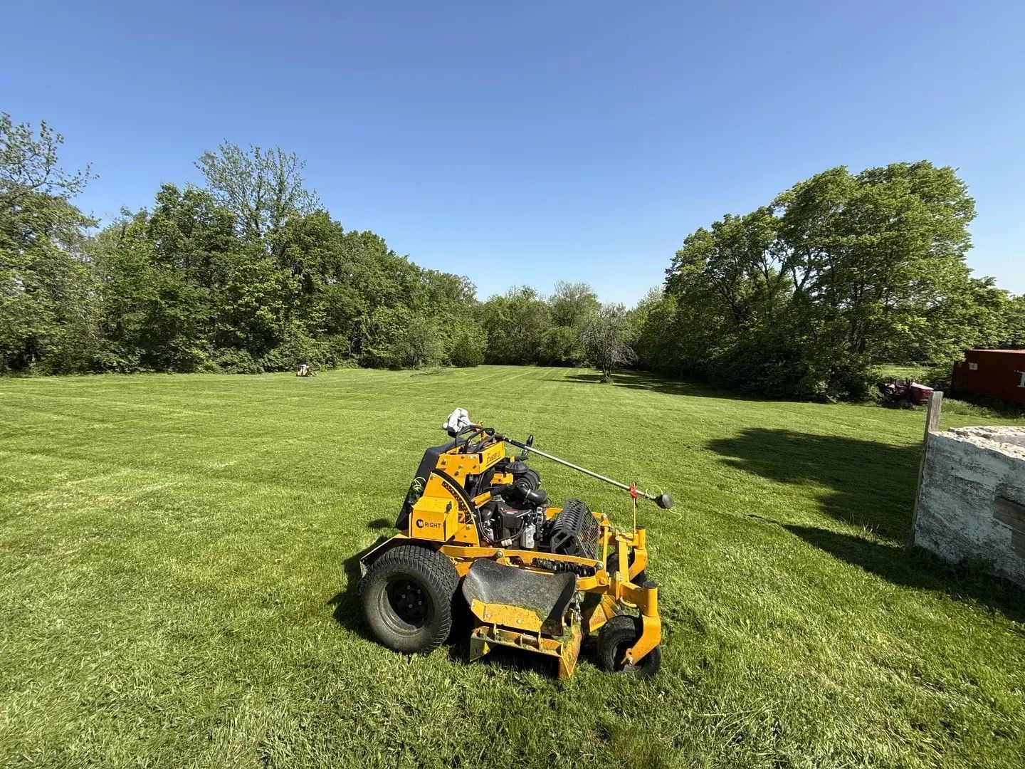 Yellow zero-turn mower on green grass in a field with trees under a blue sky.