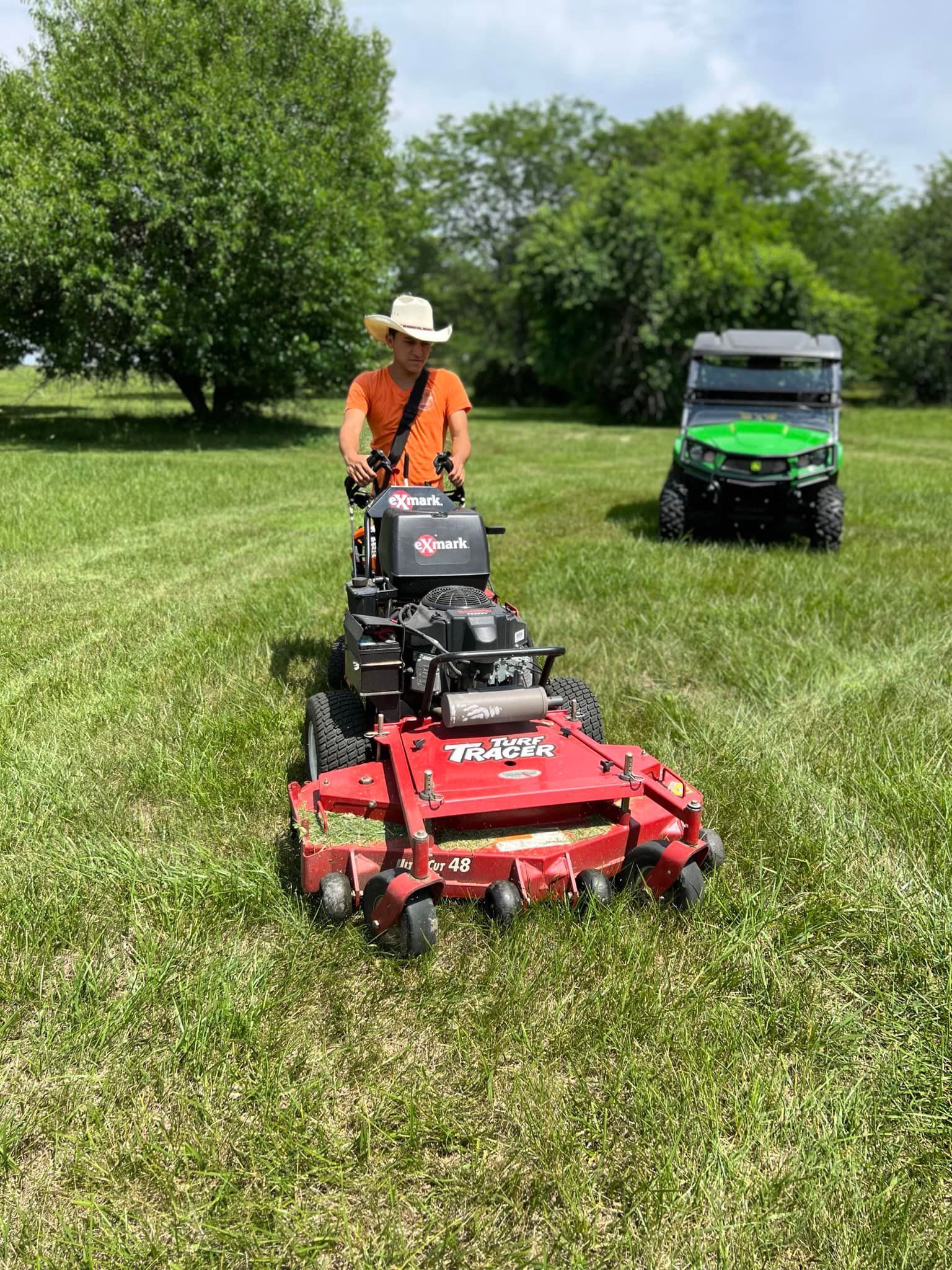 Person mowing lawn with a red zero-turn mower; a green utility vehicle is parked nearby.