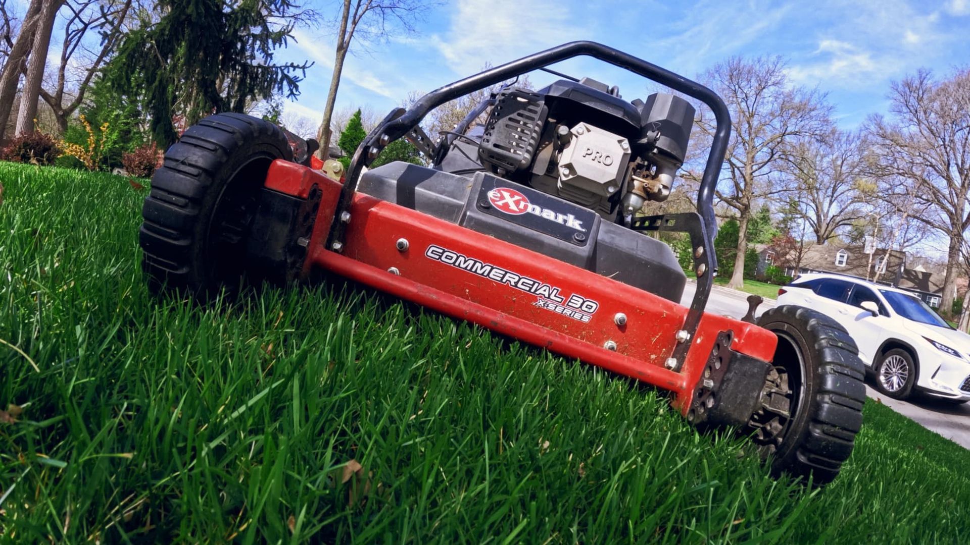 Red lawn mower cutting grass on a sloped yard, white car in background.