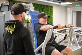 Een man zit op een apparaat in een sportschool en praat met een andere man.
