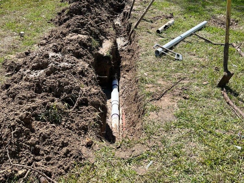 Open trench with exposed utility pipe in a grassy yard, dirt piled along the side, tools nearby.
