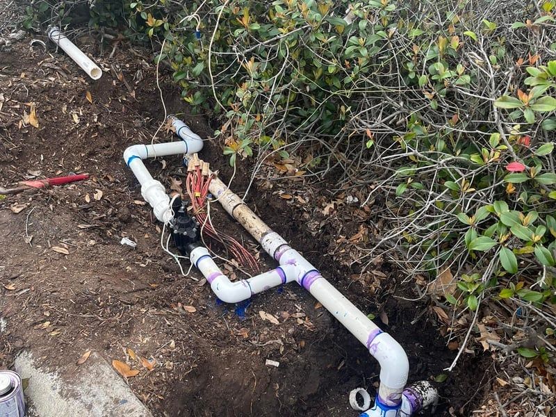 Exposed white PVC pipes with fittings along a dirt trench beside bushes