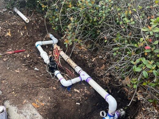 White PVC pipes and valves installed in a dirt trench beside green shrubs.