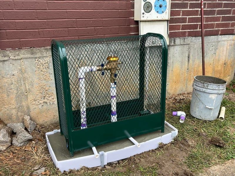 Outdoor utility gas meter enclosed in green fencing beside a brick wall and bucket